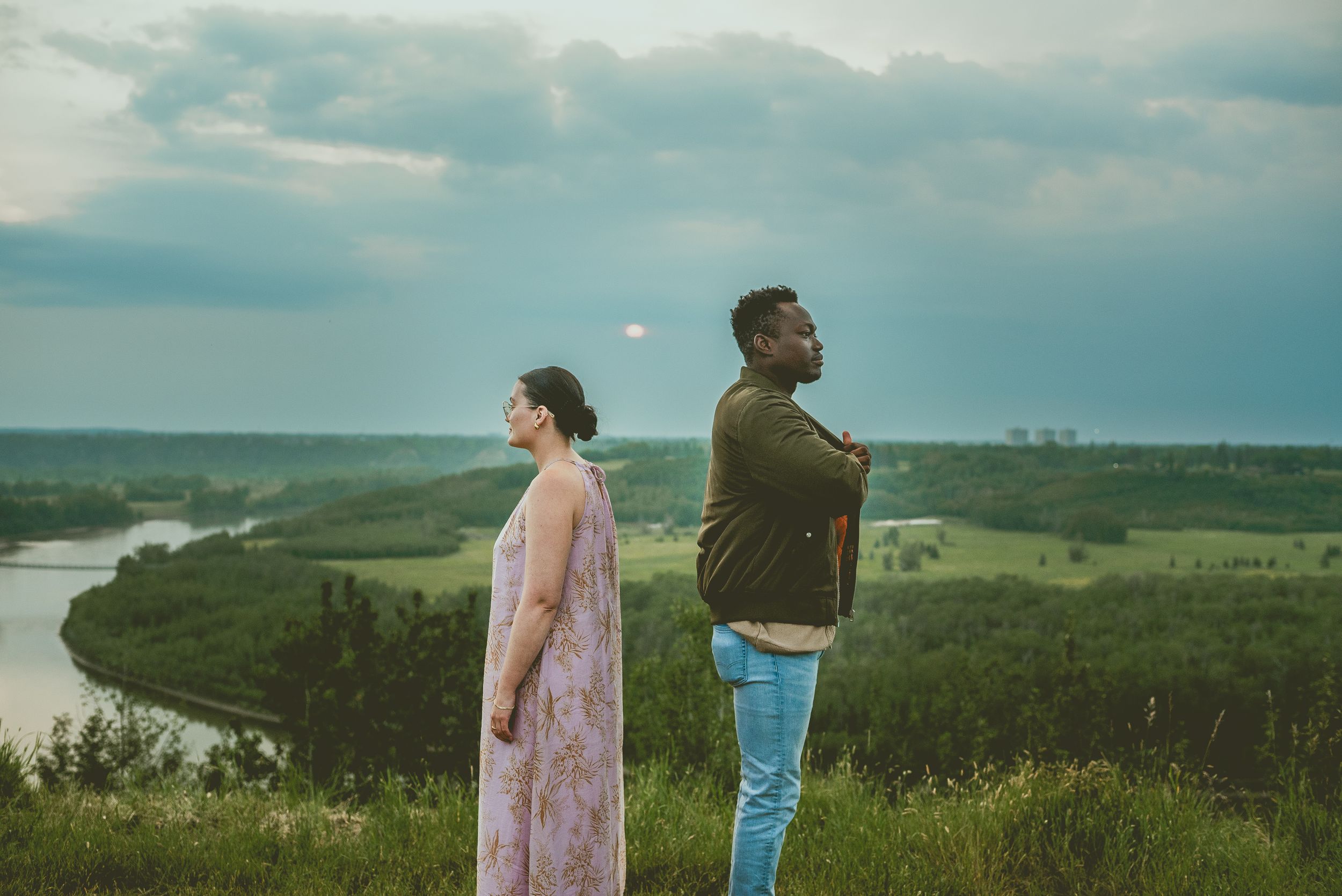 Two people stand distanced apart on a hilltop overlooking a scenic river valley landscape at dusk.