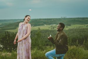 Romantic proposal scene on a grassy hilltop with rolling green countryside landscape in the background during sunset.