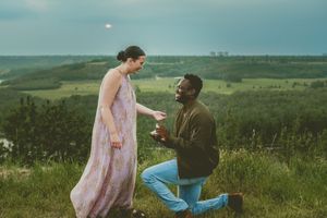 A proposal moment unfolds on a grassy hillside as two people share a romantic moment at sunset.