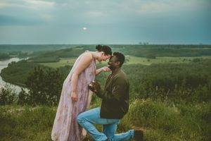 A romantic proposal scene overlooking a winding river valley with rolling hills and misty sky at sunset.