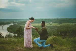 People sit together on a scenic hilltop overlooking a vast green countryside landscape with rolling hills and grass fields.