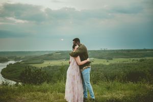 Two people embrace while standing on a green hilltop overlooking a scenic rural landscape under moody skies.