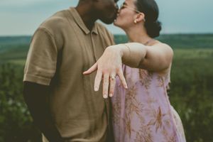 Engaged couple kiss in a field while showing off a new engagement ring on the woman's outstretched hand.