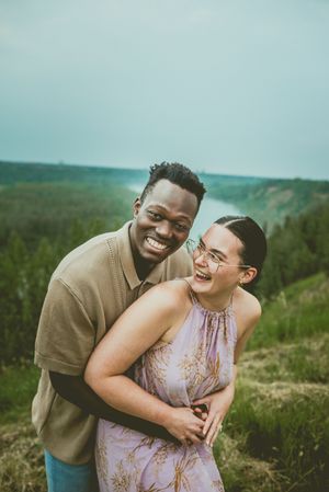 A joyful couple laughs together while embracing on a hillside with scenic green rolling landscape in the background.