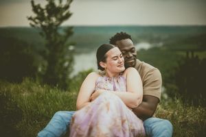 Couple shares a tender moment sitting together on a grassy hillside overlooking a scenic landscape.