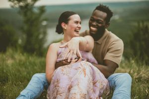 Happy couple laughs together while sitting on grass with scenic rolling hills in the background.
