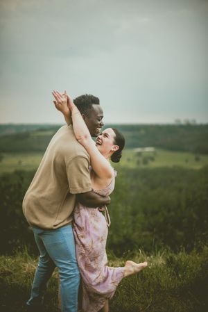 A joyful couple embraces and dances together on a hilltop overlooking green rolling countryside at sunset.