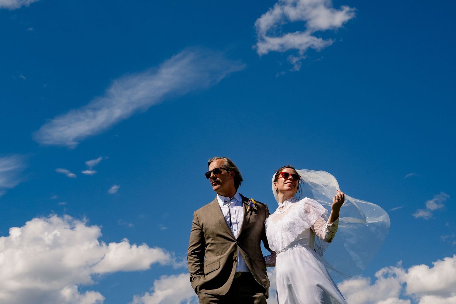 A couple poses outdoors against a bright blue sky with dramatic white clouds during their wedding celebration.