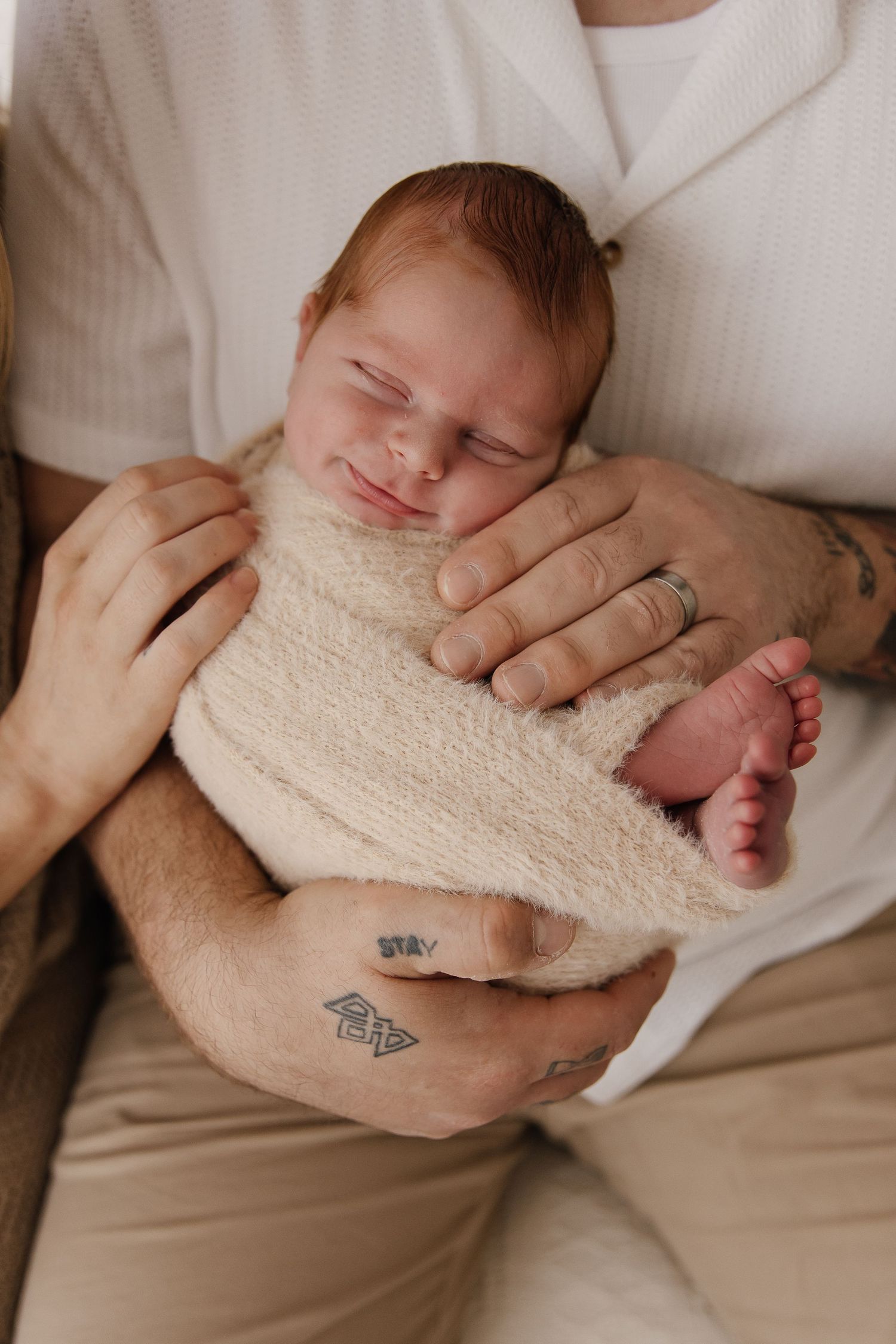 A sleeping newborn wrapped in a cream colored blanket is held gently in tattooed hands while resting peacefully.