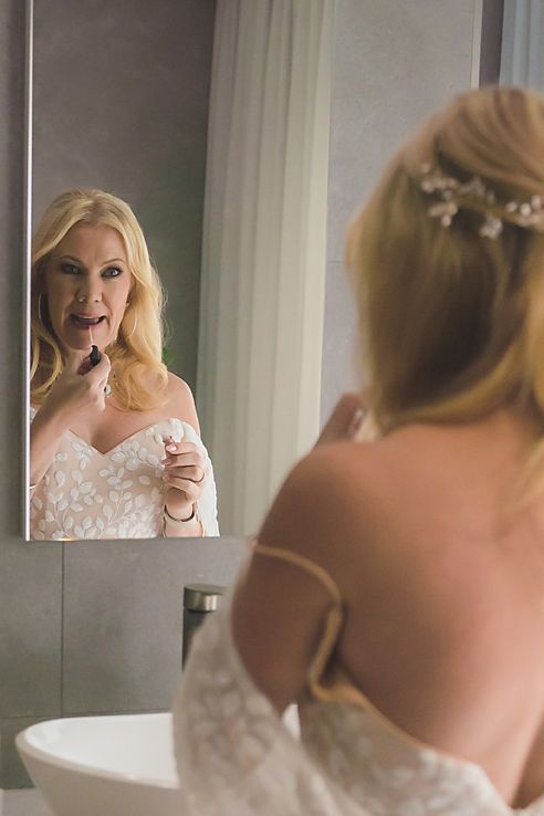 A bride in a white lace wedding dress looks in the bathroom mirror while applying lip product before her ceremony.