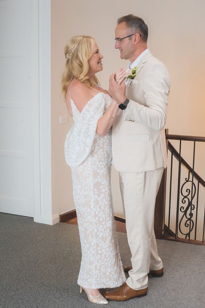 A couple shares an intimate dance moment in formalwear, with the woman in a white patterned dress and man in light attire.