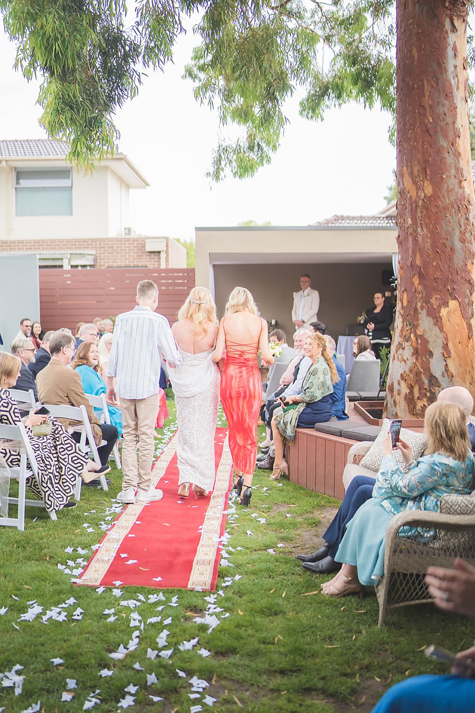 Outdoor backyard wedding ceremony with guests seated on lawn chairs along a red carpet aisle under eucalyptus trees.