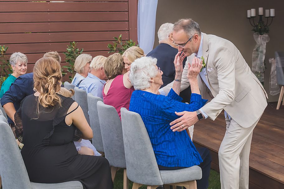 Wedding guests seated in chairs interact with person in beige suit during an indoor ceremony.