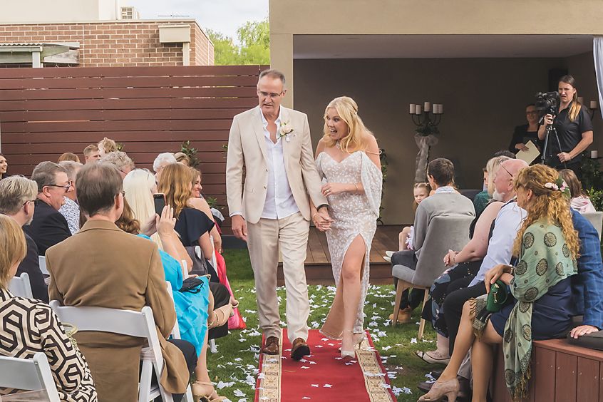 Wedding ceremony with guests seated outdoors on chairs along a red carpet aisle during a daytime celebration.