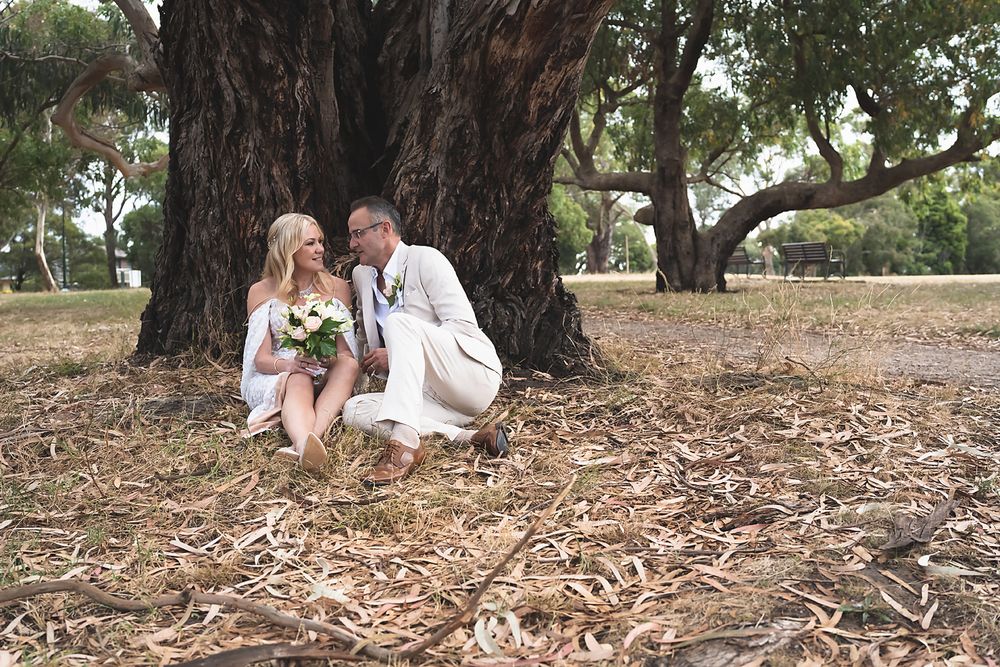 A couple in wedding attire sits together under a large tree trunk on dried leaves in a park setting.