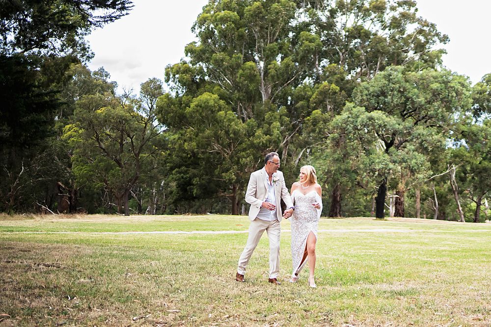 A couple walks together holding hands on a sunny day in a grassy park with tall eucalyptus trees in the background.
