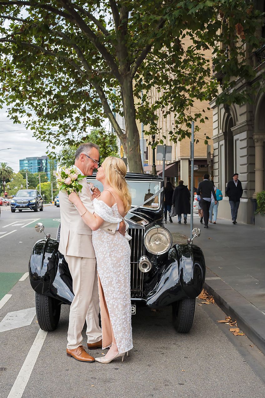 A vintage car photoshoot featuring a romantic moment between a couple in formal attire on a tree-lined street.