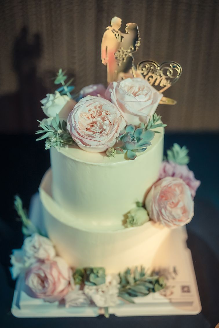 A two-tiered white wedding cake decorated with pale pink roses, succulents, and a gold love topper.