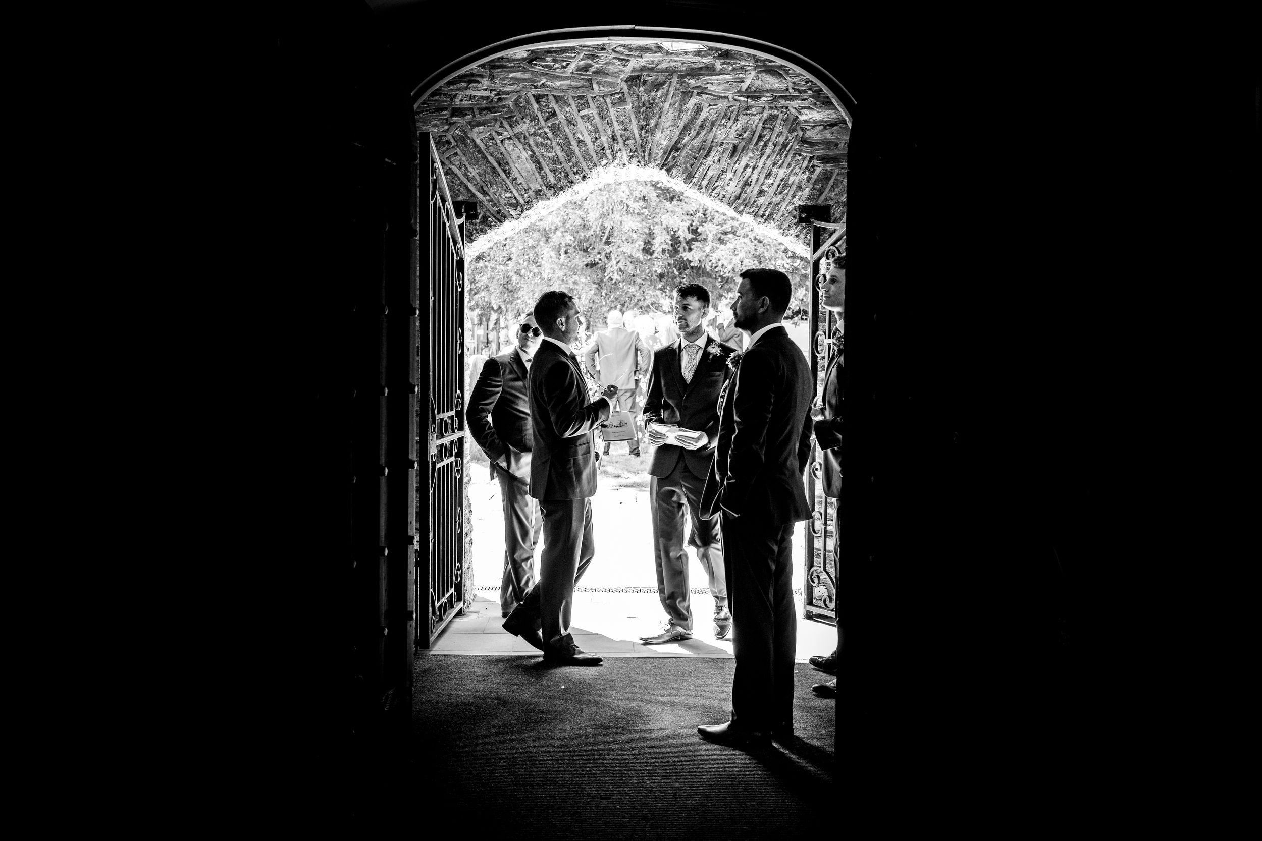 Black and white silhouette of three groomsmen conversing in an arched doorway before a wedding.