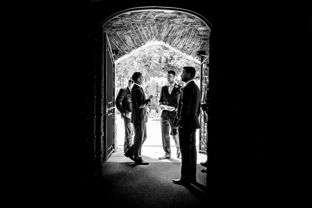Black and white silhouette of three groomsmen conversing in an arched doorway before a wedding.