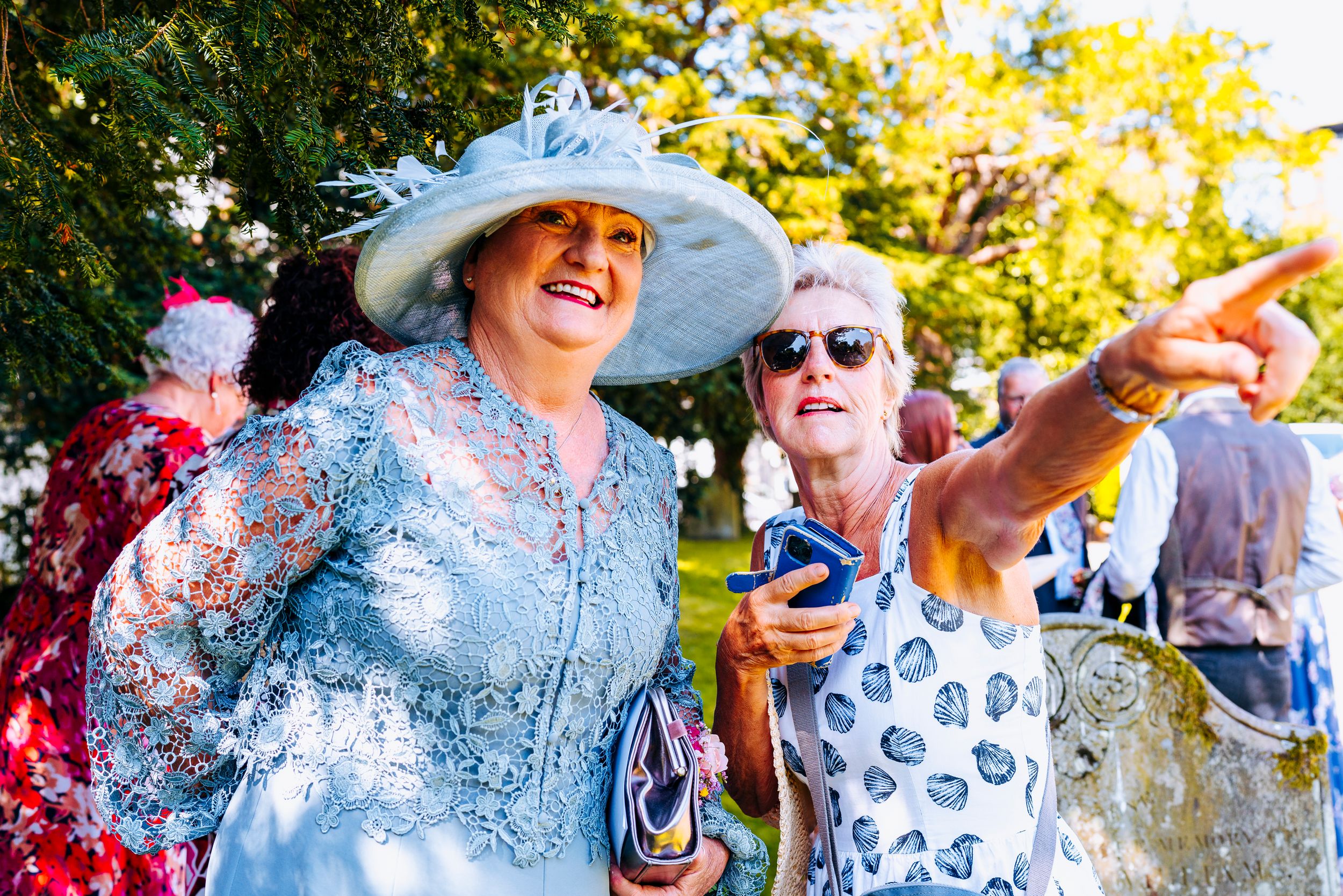 Two elegantly dressed wedding guests wearing hats smiling and pointing outdoors at a wedding.