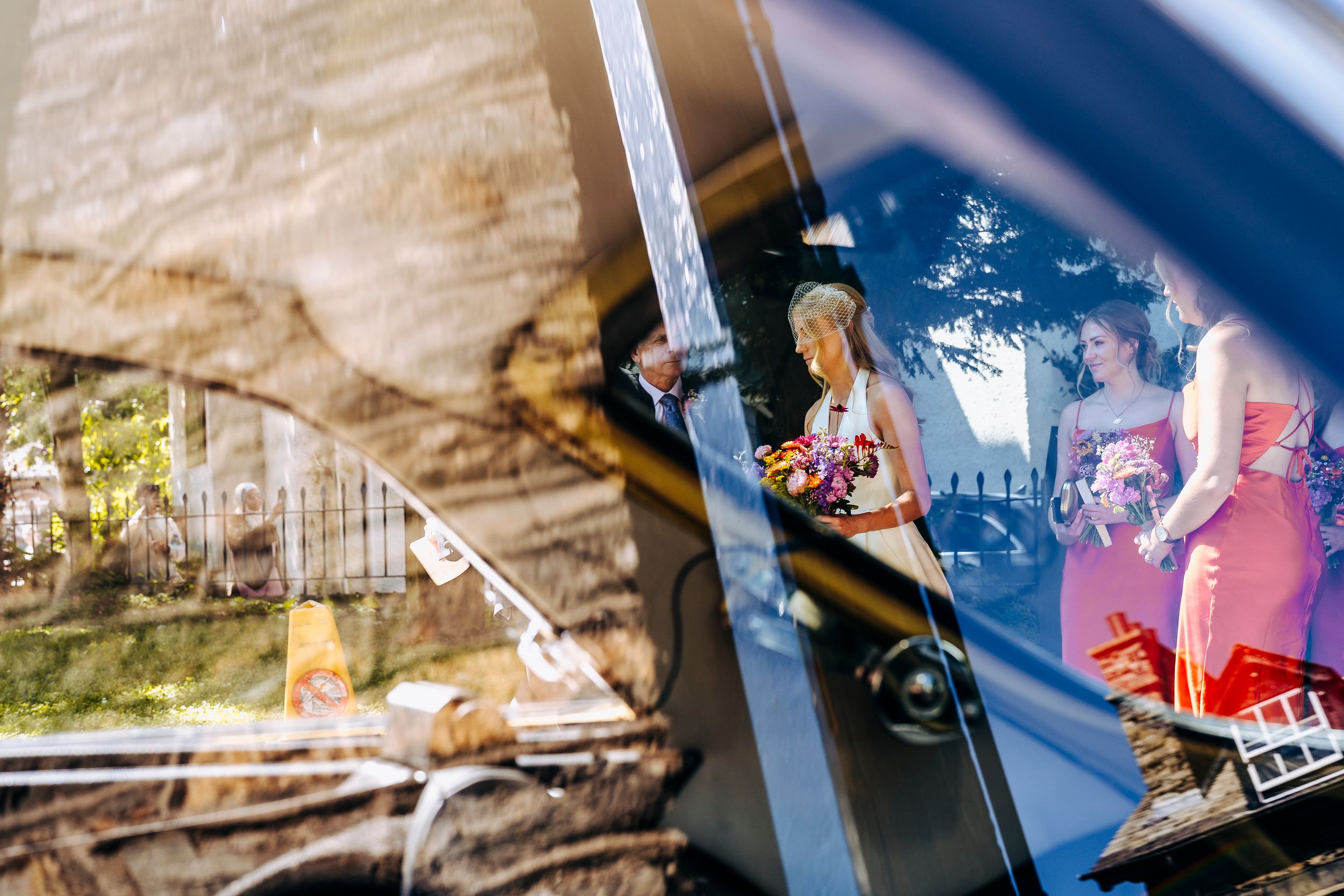 Creative car window reflection capturing bride with bouquet and bridesmaids outside the church.