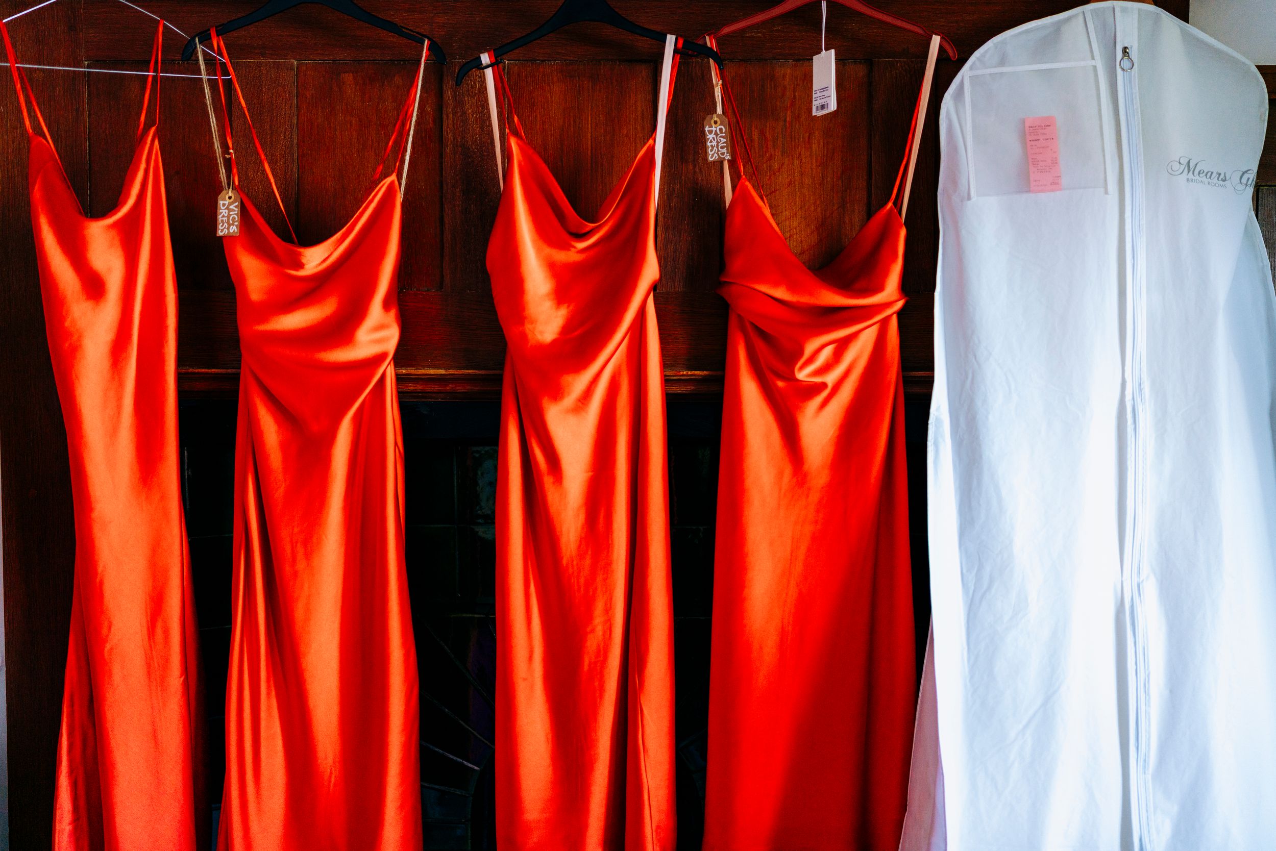 Three red satin bridesmaid dresses hanging beside a white bridal gown in a garment bag.