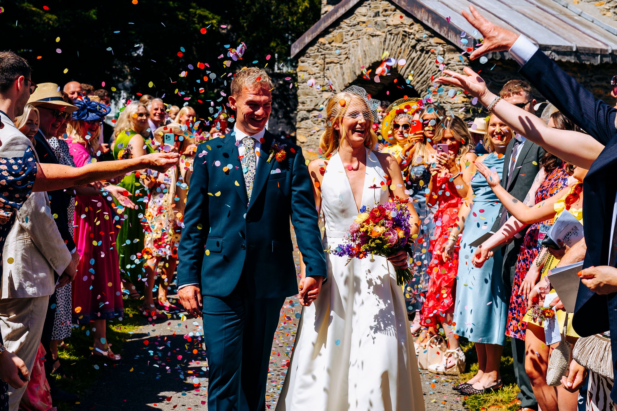 Bride and groom walk through colorful confetti shower outside stone church on sunny wedding day.