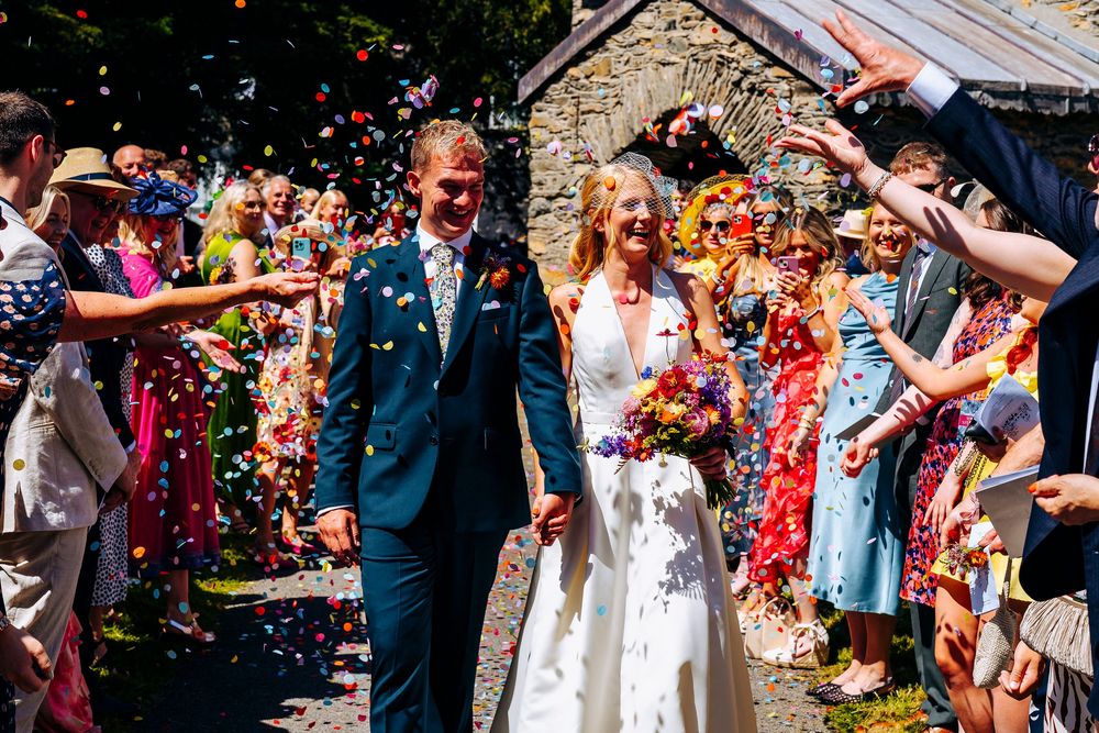 Bride and groom walk through colorful confetti shower outside stone church on sunny wedding day.