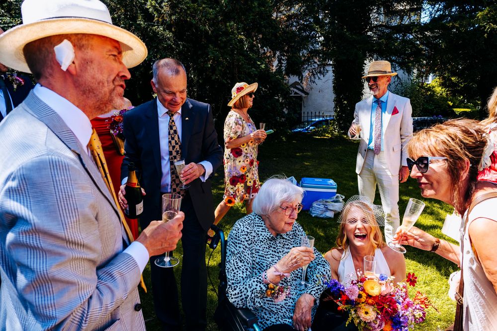 Wedding guests laugh and socialize outdoors in summer sunshine wearing elegant hats and attire.