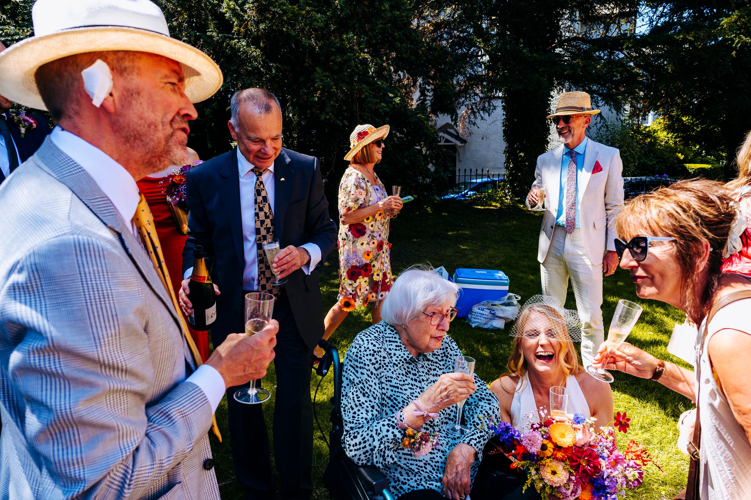 Wedding guests laugh and socialize outdoors in summer sunshine wearing elegant hats and attire.