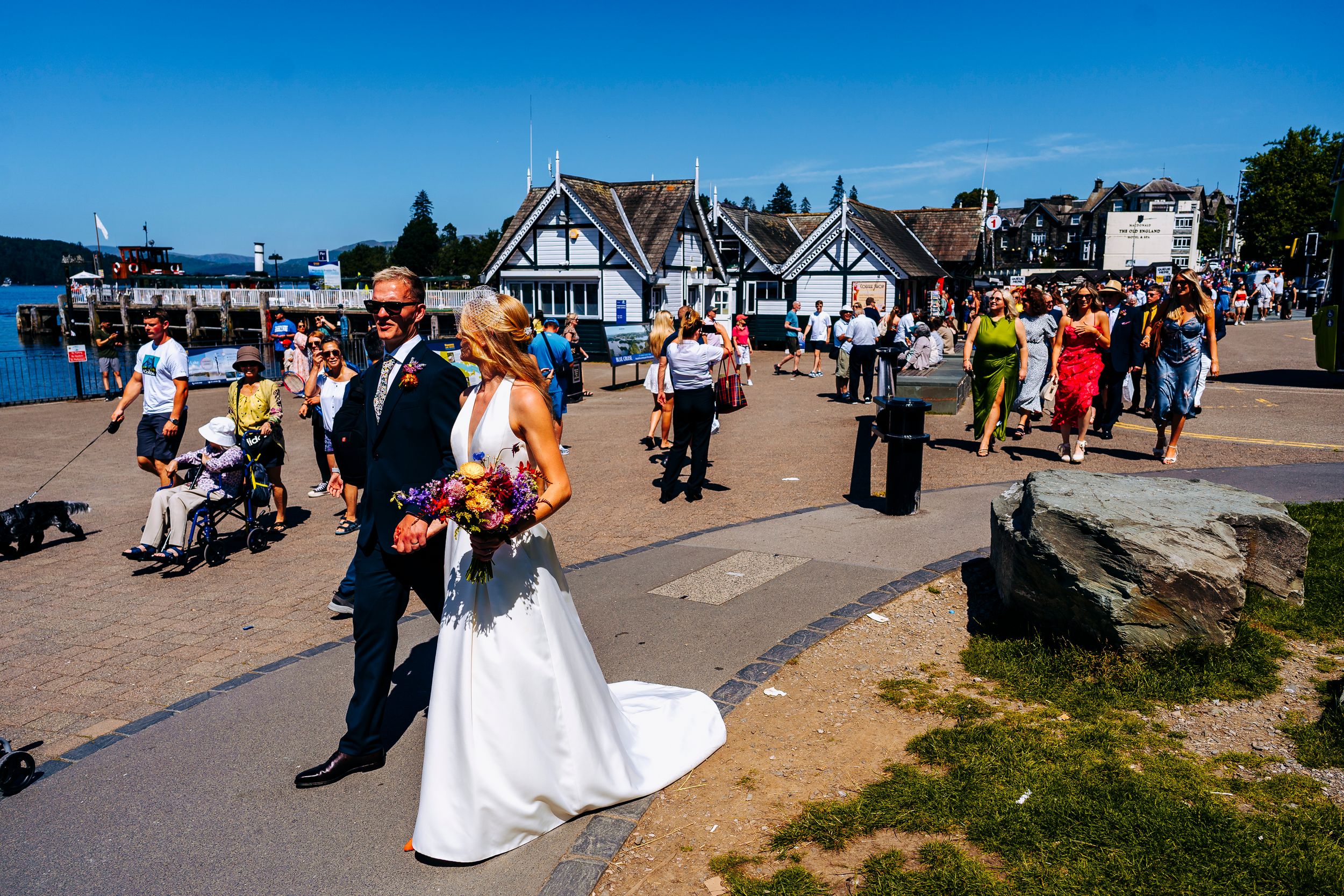 Bride in white gown and groom walk along sunny lakeside promenade with wedding guests nearby.
