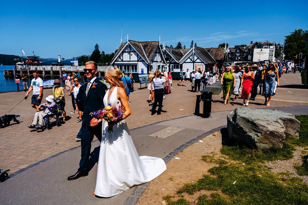 Bride in white gown and groom walk along sunny lakeside promenade with wedding guests nearby.