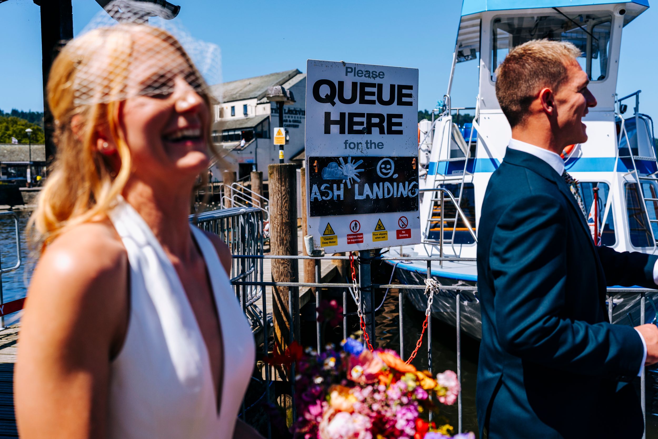 Laughing bride in white dress stands near queue sign at sunny lakeside boat dock with groom.