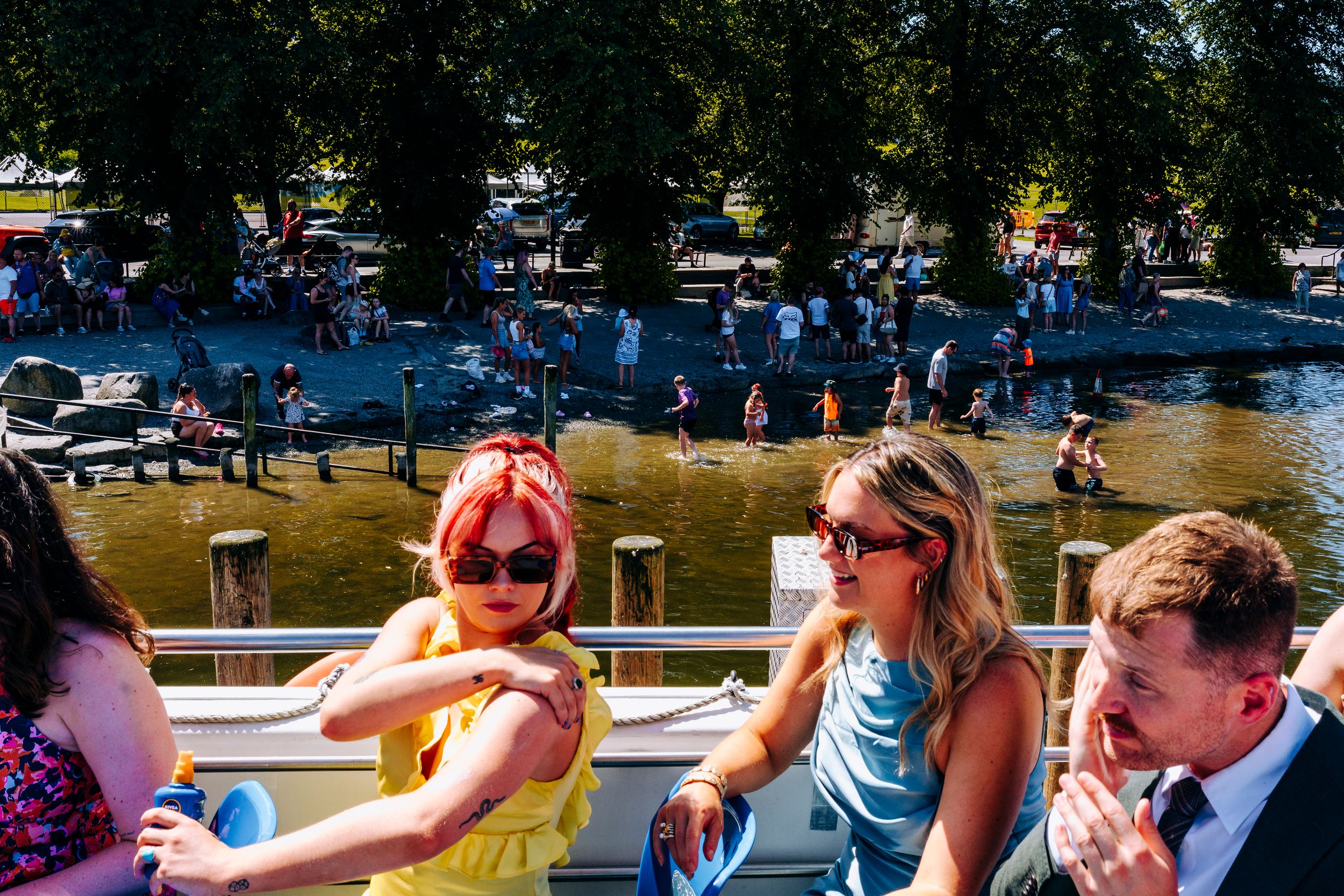 Wedding guests enjoy sunny boat ride on lake with woman in yellow dress wearing red sunglasses.