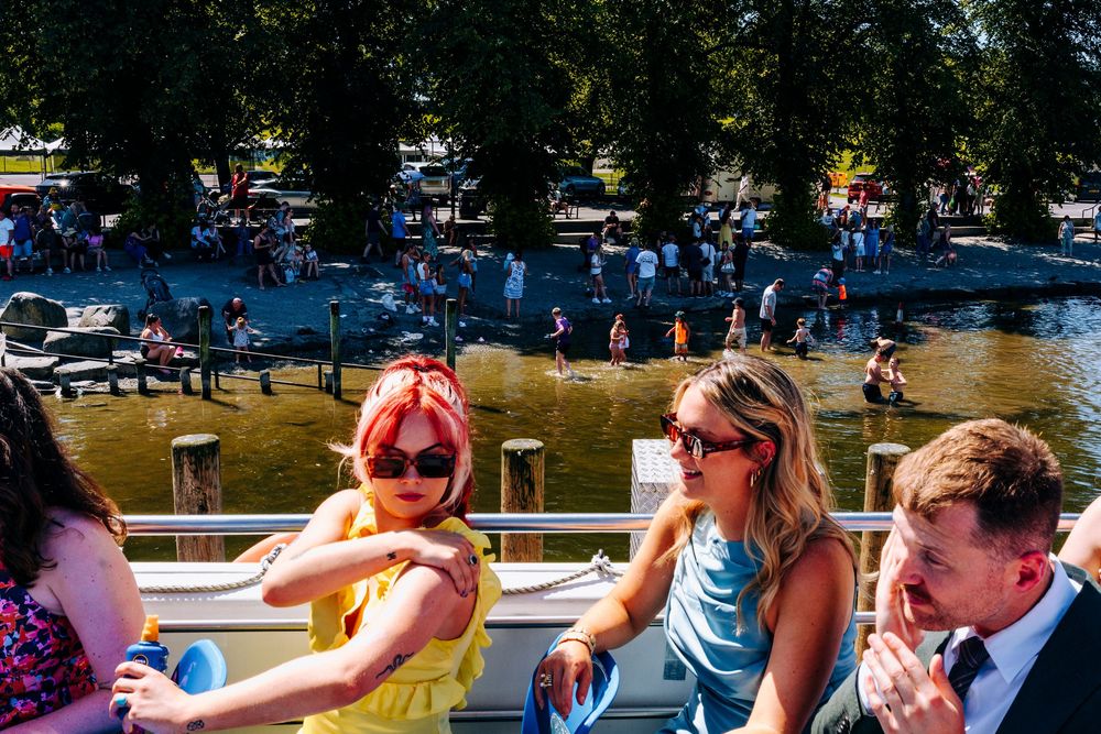 Wedding guests enjoy sunny boat ride on lake with woman in yellow dress wearing red sunglasses.
