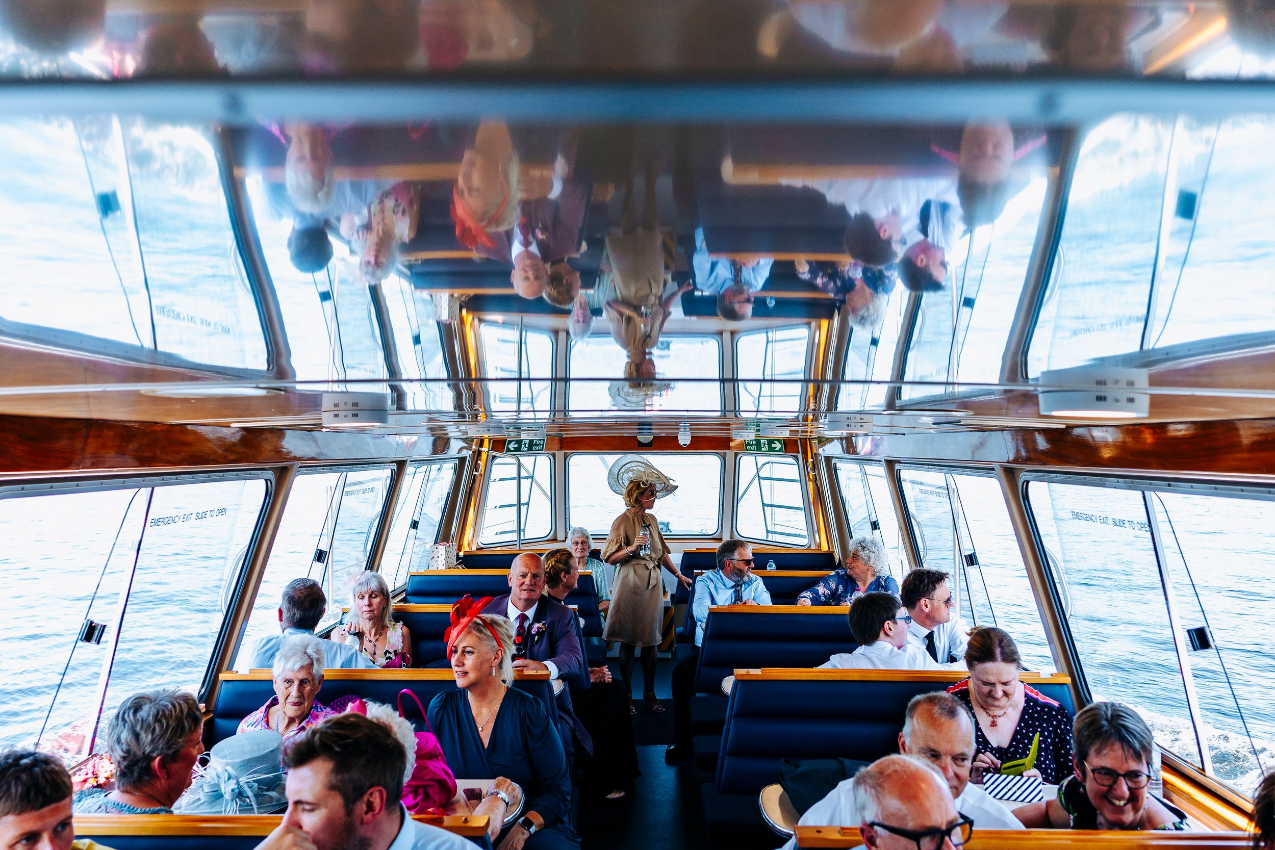 Wedding guests seated inside glass-roofed passenger boat cruising on sparkling water on sunny day.
