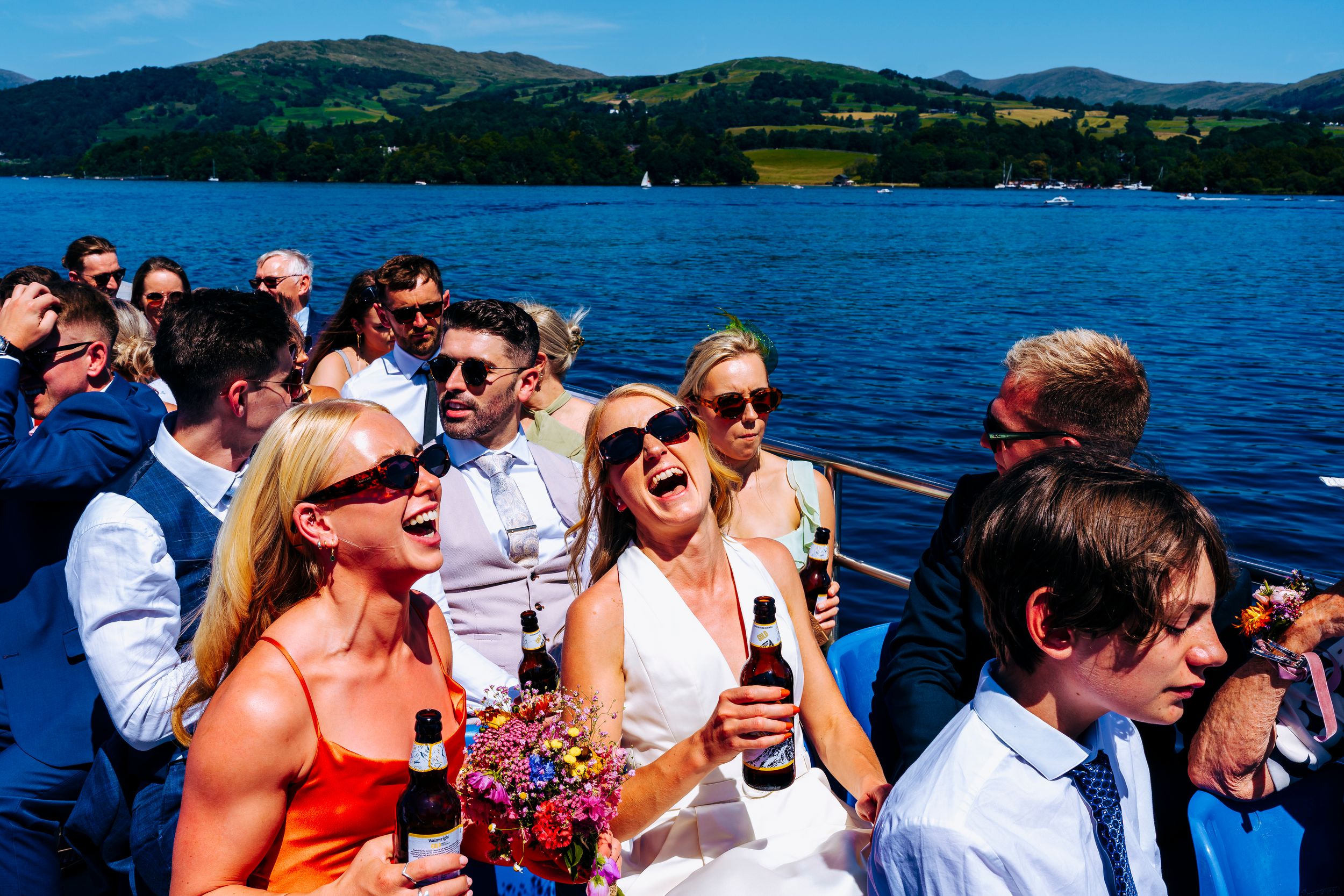 Wedding guests laughing and celebrating with beers on a boat on a scenic lake Winderere.