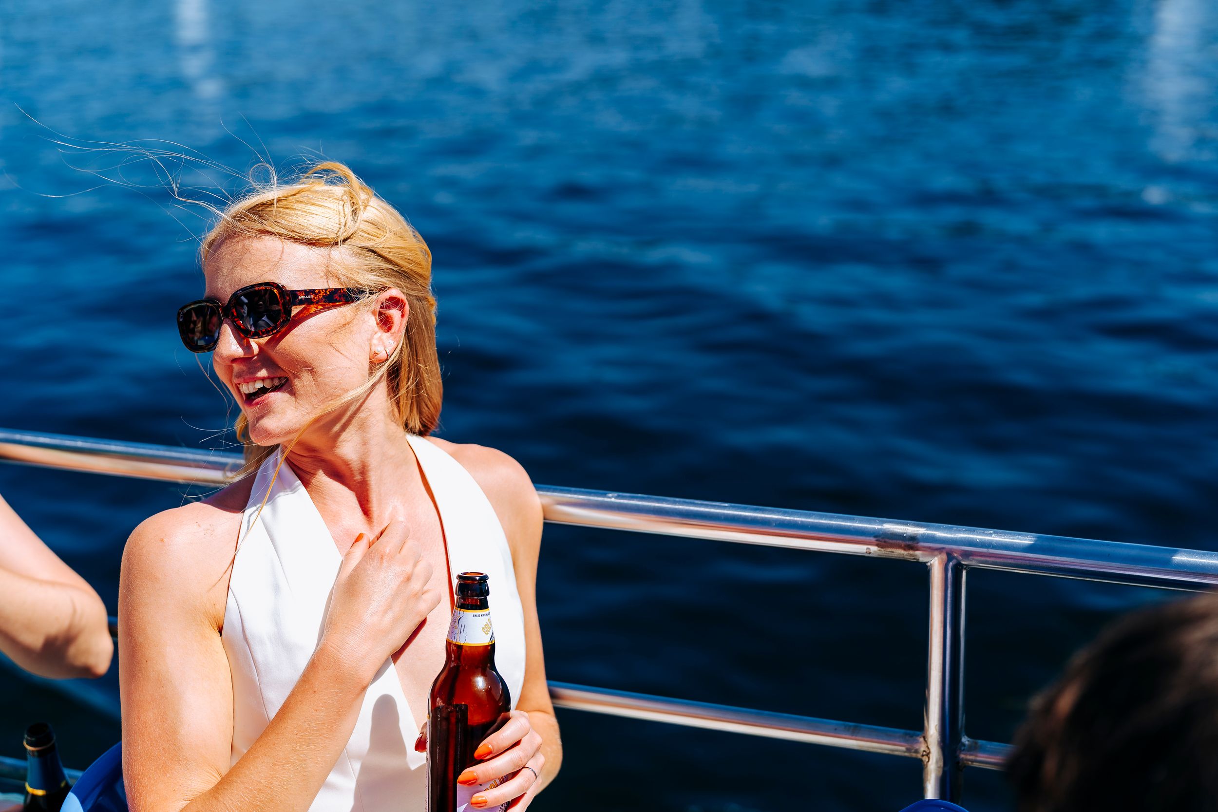 Bride in white dress holding a beer bottle on a sunny boat on blue water of Lake Windermere