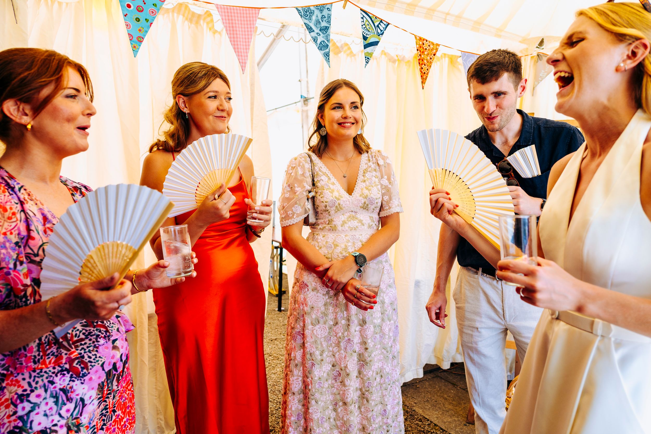 Wedding guests using hand fans to cool down inside a decorated marquee tent.