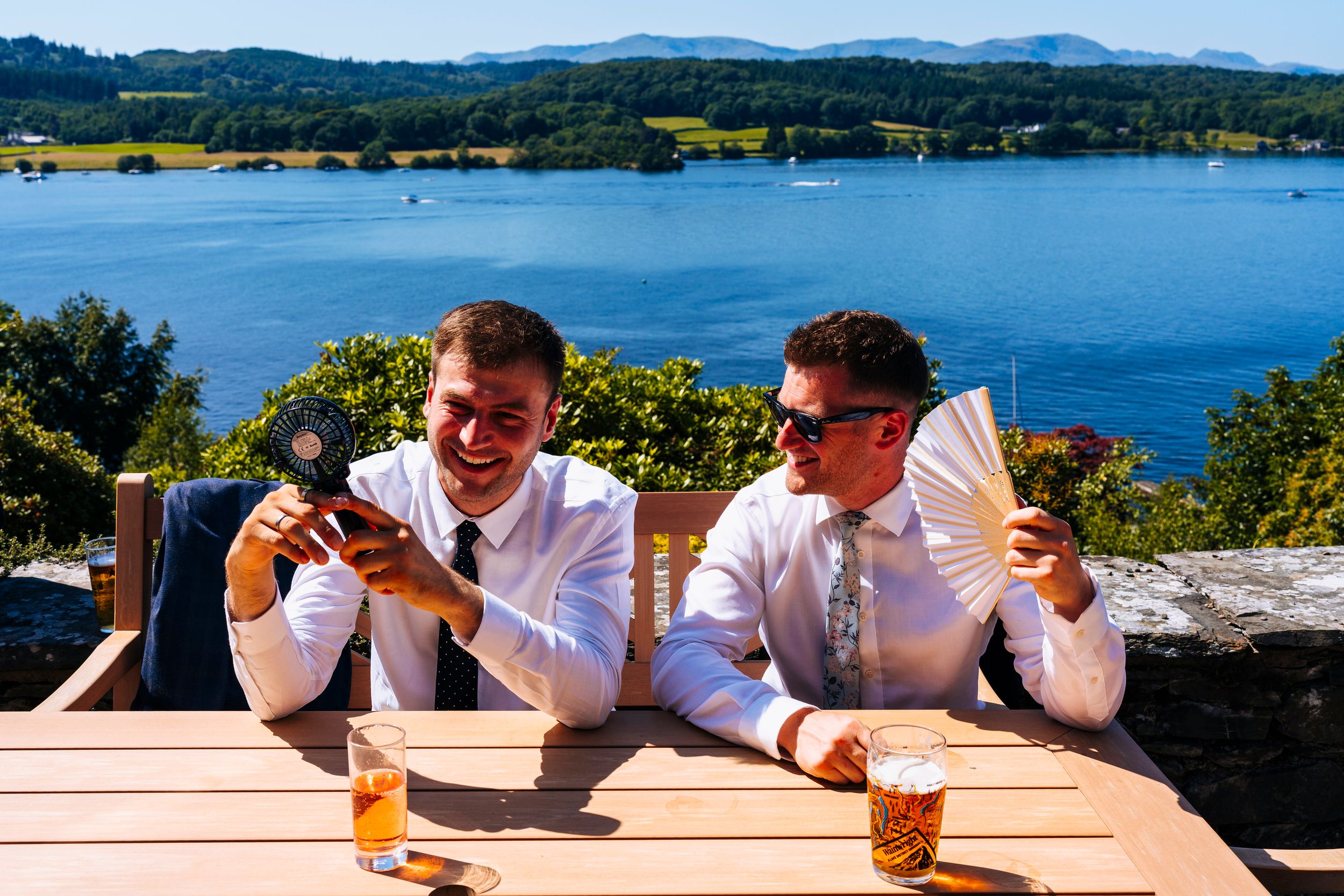 Two men laughing and using hand fans at an outdoor table overlooking a lake.