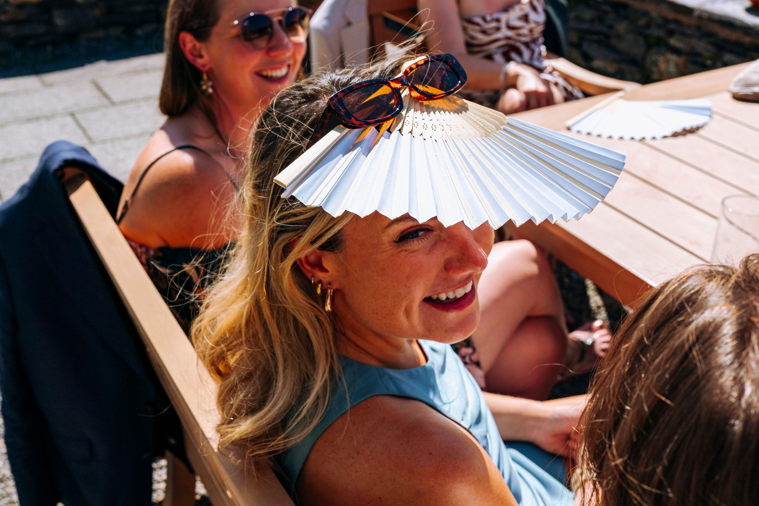 Woman laughing and using a white hand fan as a sun shield at an outdoor event.