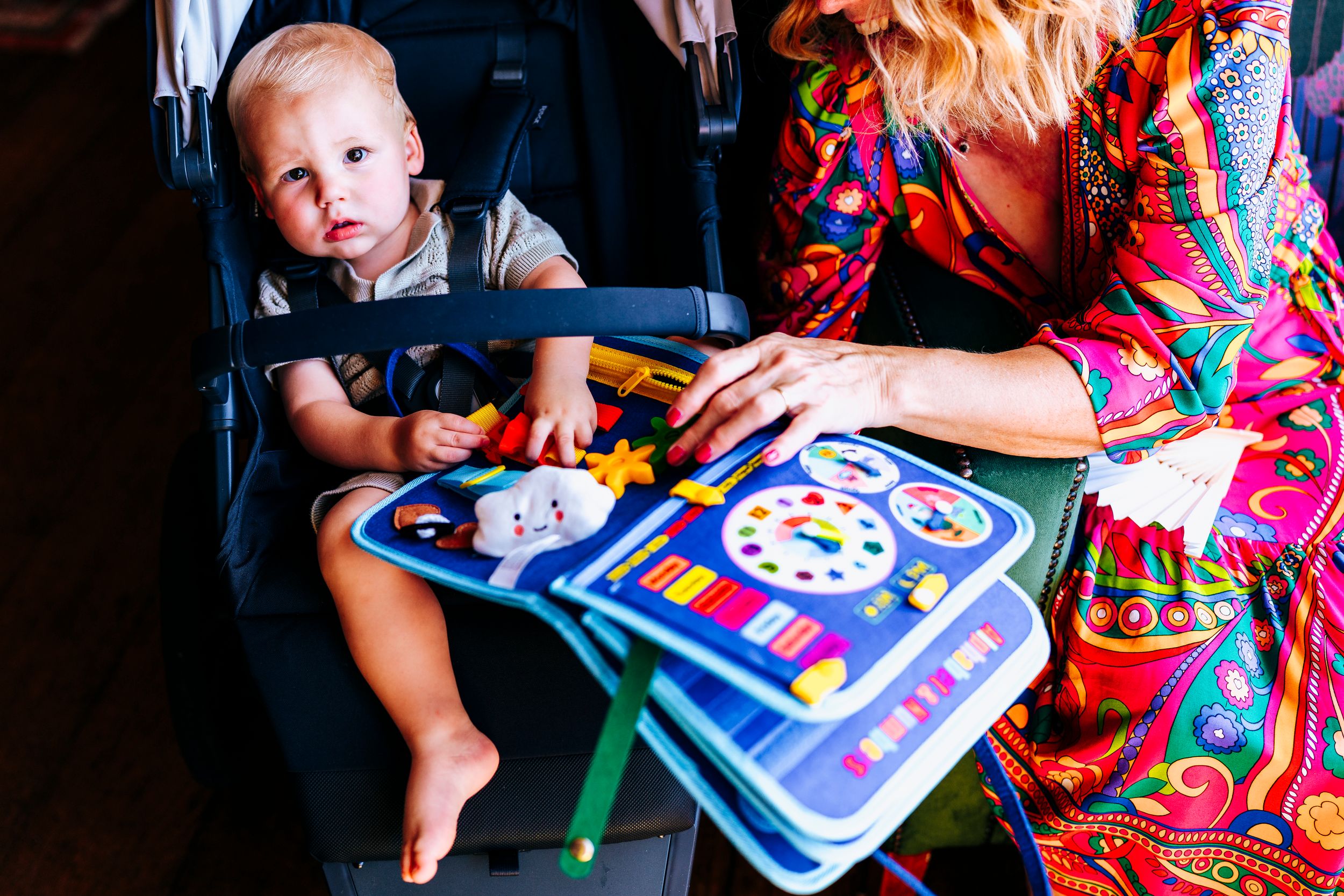A mother in a colorful dress shows her toddler an activity board while the baby sits in a car seat.