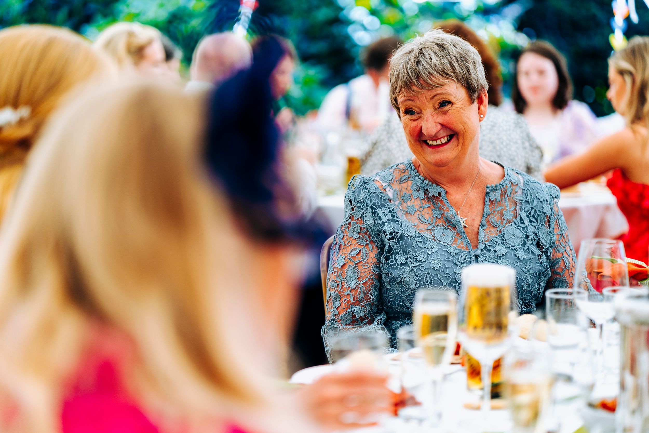 An elegantly dressed older woman smiles warmly at a wedding reception table with champagne glasses.