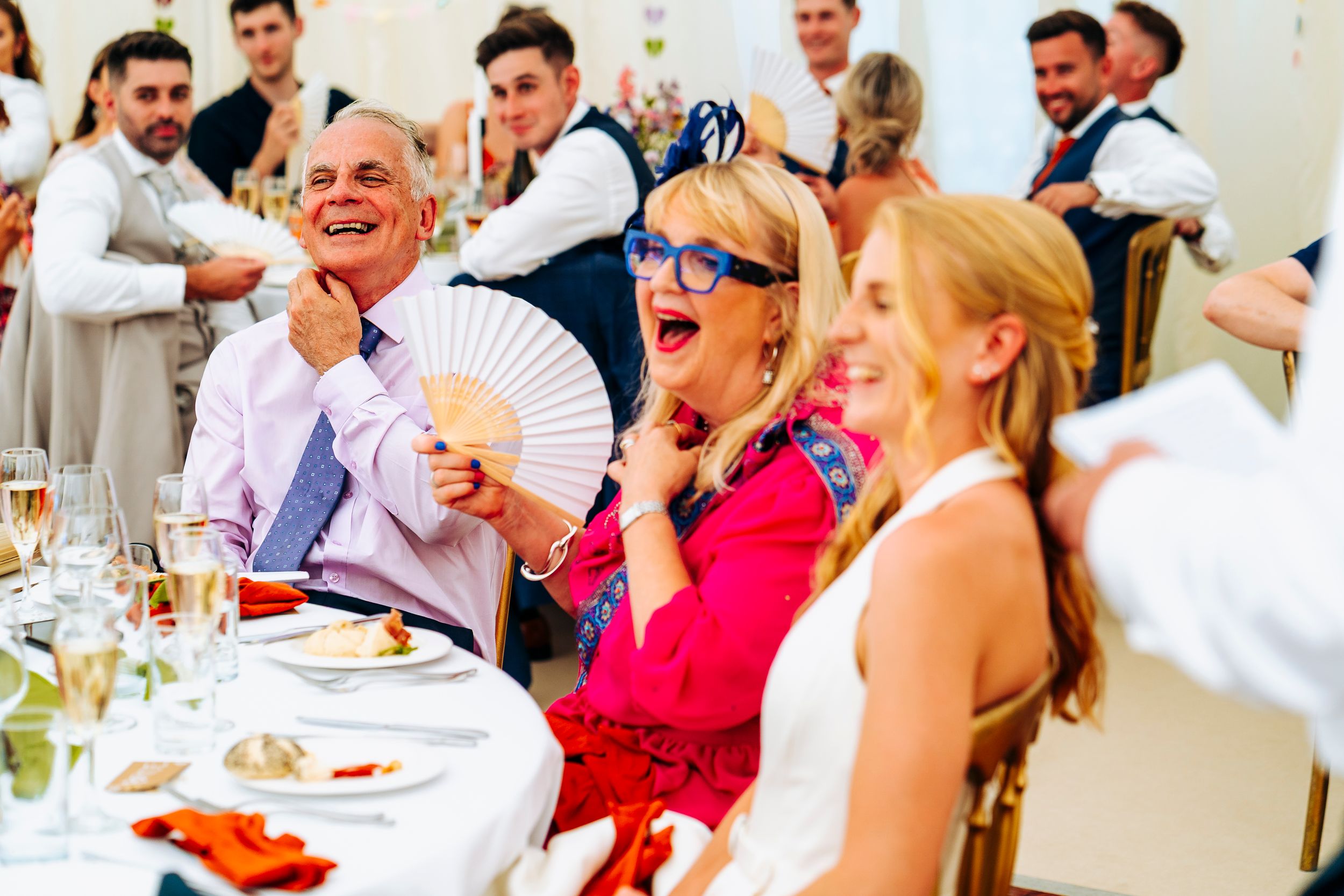 Wedding guests laugh and fan themselves at a sunny outdoor reception under a white marquee tent.