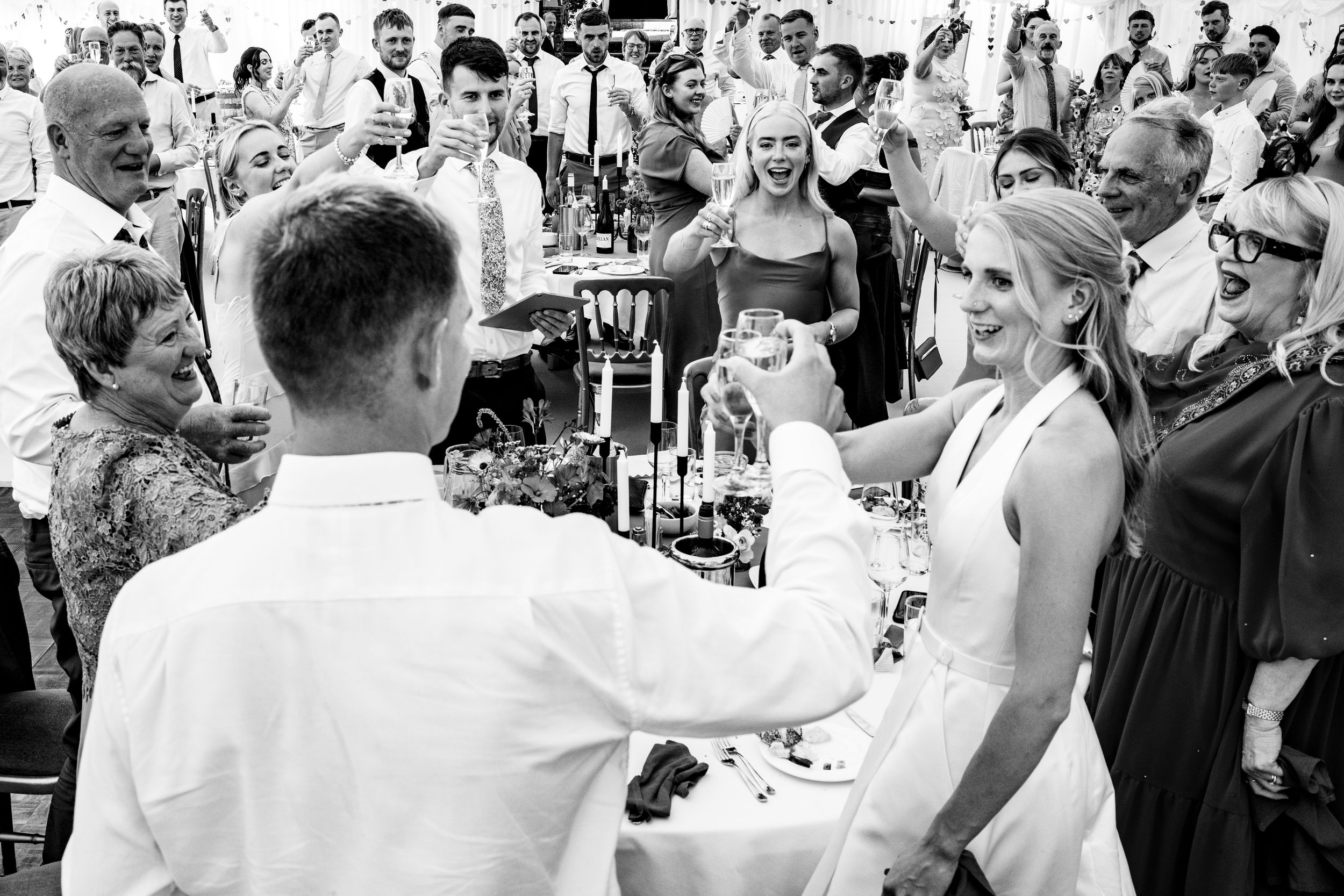 A bride and groom share a champagne toast surrounded by cheering guests at their wedding reception.