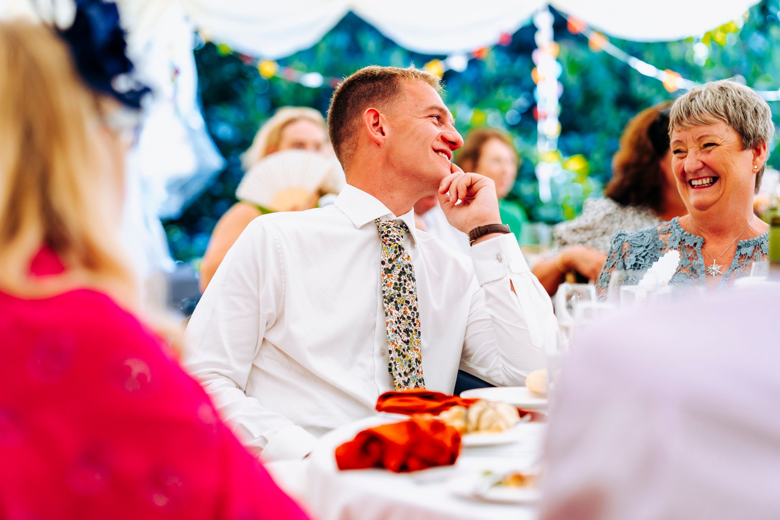 A smiling man in a floral tie enjoys a laugh with guests at a wedding reception.