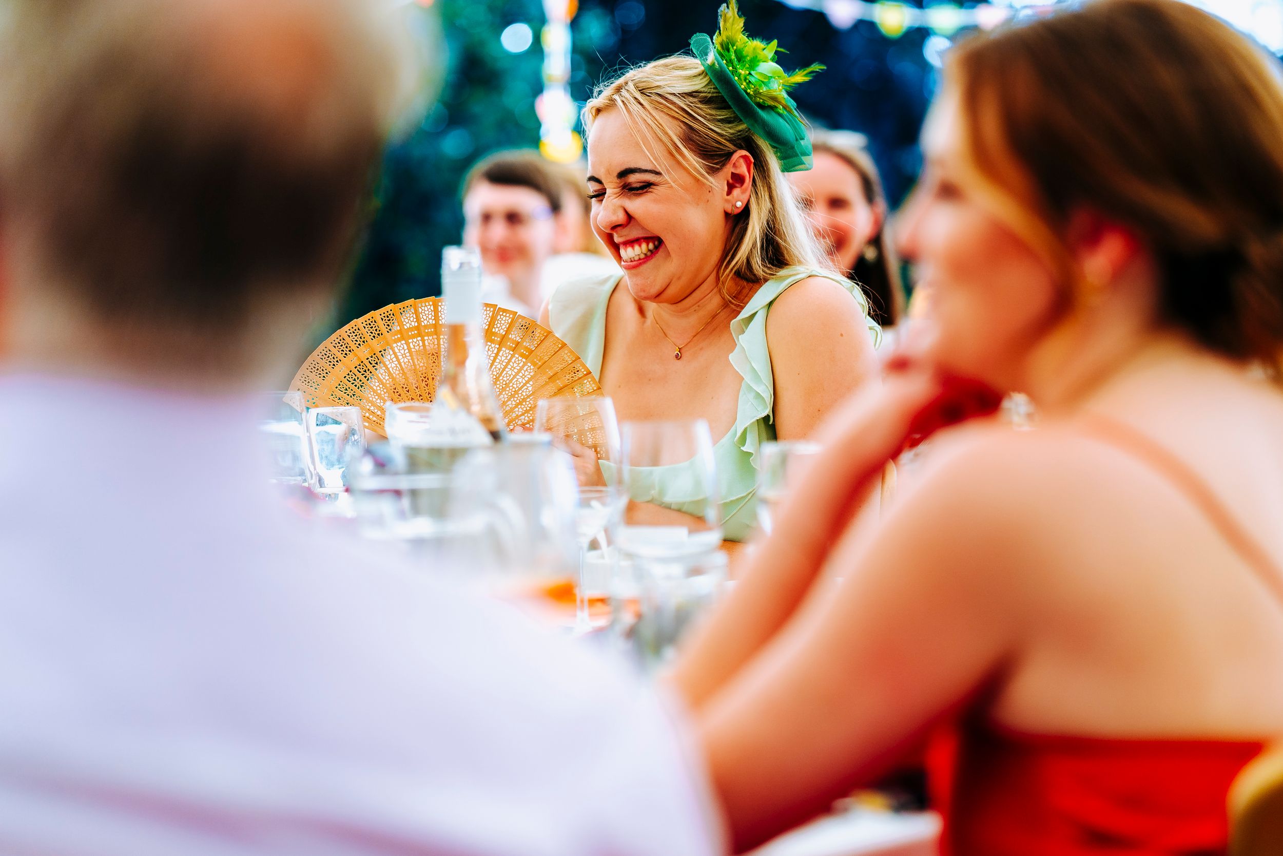 A joyful wedding guest in a green headpiece laughs at the reception dinner table.