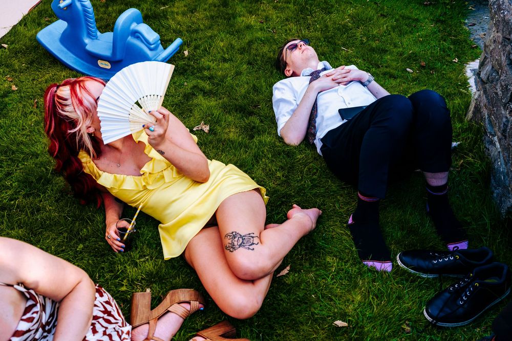Women relaxing on grass at outdoor wedding, one in yellow dress holding fan, another with tattoos resting.