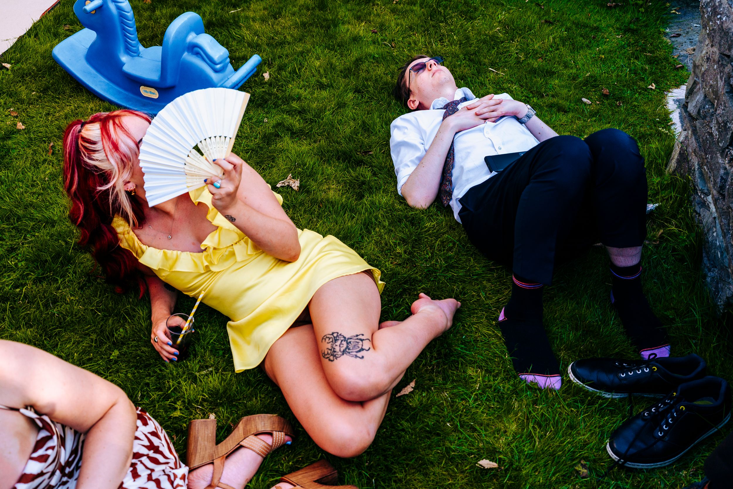Women relaxing on grass at outdoor wedding, one in yellow dress holding fan, another with tattoos resting.